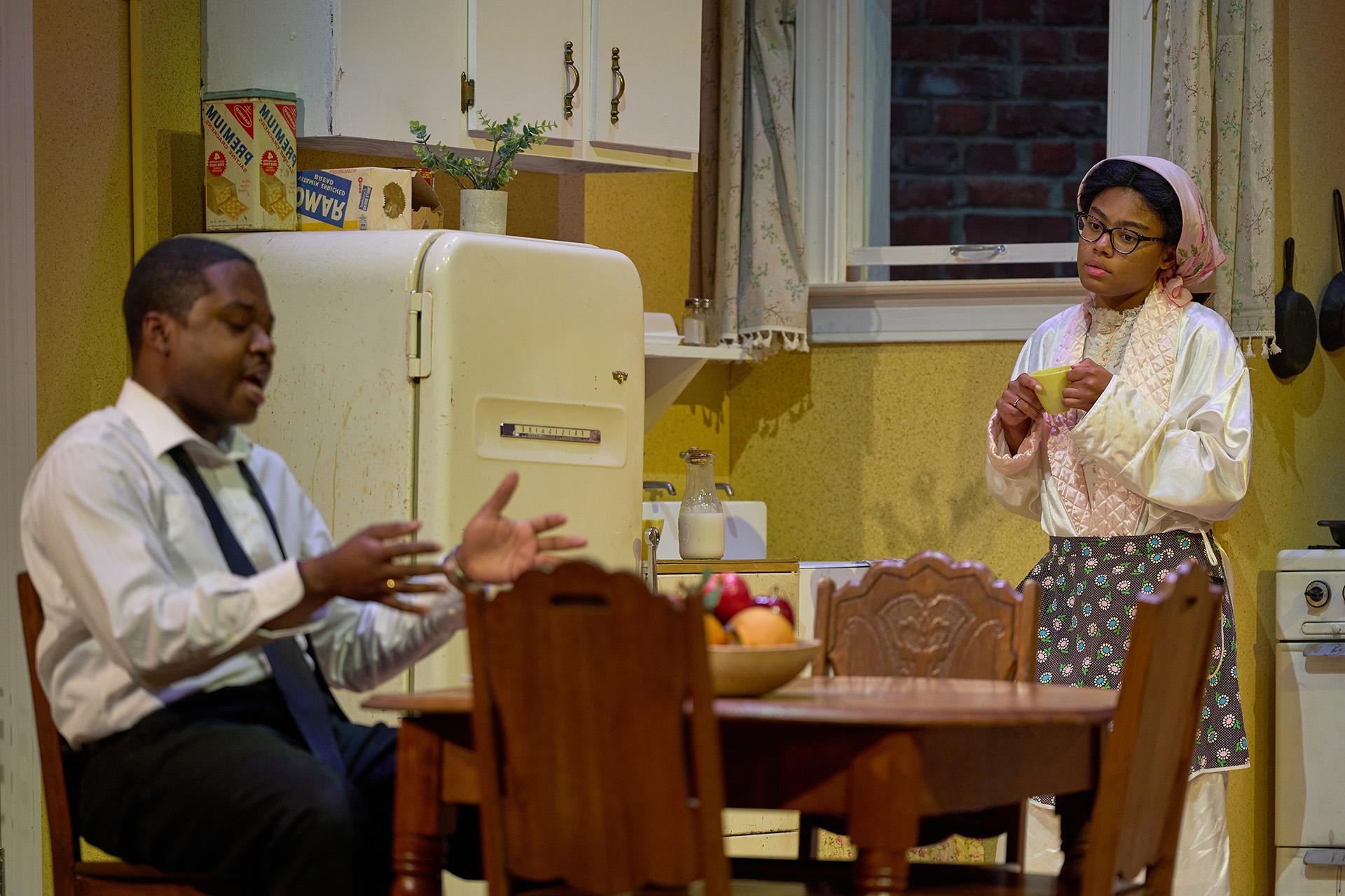 A young Black woman in houseclothes holds a teacup while looking at her husband seated at the kitchen table